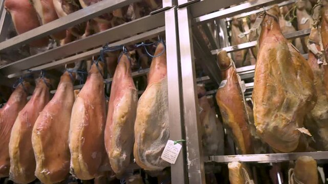 Smooth camera pan of authentic Spanish hams with Protected Designation of Origin tags aging on stainless steel racks inside an industrial curing facility