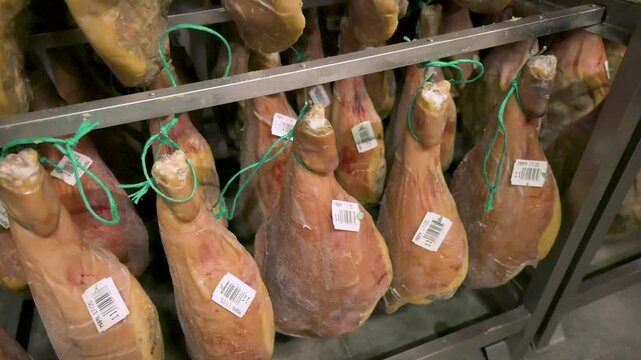 Smooth camera pan of authentic Spanish hams with tracking barcode tags aging inside an industrial curing room or drying cellar facility