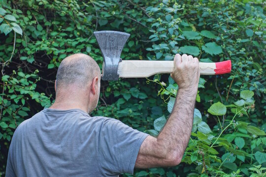 one male criminal in gray clothes  swings hands a big axe aggressively on the street among the trees