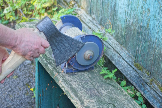 One male worker holds a large axe in his hands and sharpens the iron blade on a grinding wheel on an electric sharpening machine on a table