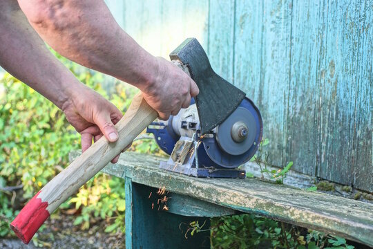 One male worker holds a large axe in his hands and sharpens the iron blade on a grinding wheel on an electric sharpening machine on a table