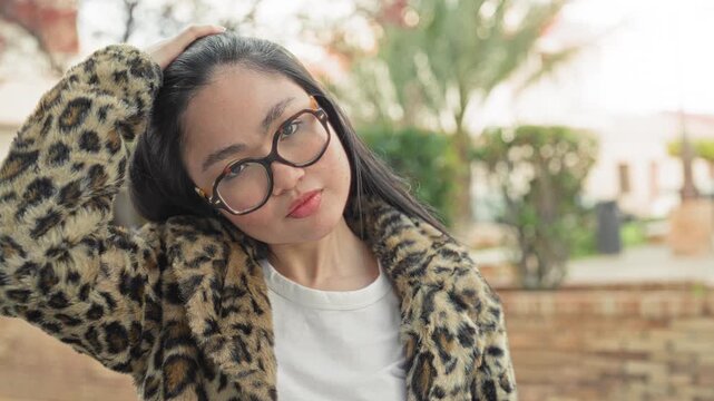 Woman with tortoiseshell glasses wearing a leopard coat touches hair and smiles on a street beside a brick wall; contentment charm.