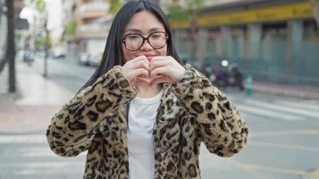 Woman smiling with eyes closed makes heart shape with hands on street wearing leopard coat and glasses, near crosswalk and scooters; joy affection.