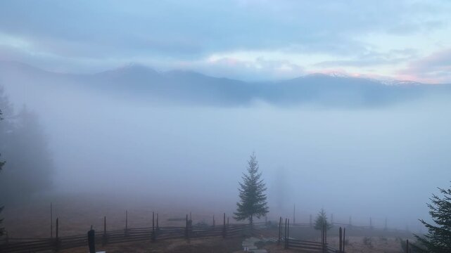 Misty mountain pasture at dawn with quiet forest and distant peaks, soft video frame showing fog drifting over wooden fences, trees and blue ridges