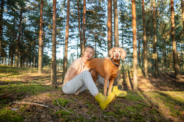Fototapeta premium Thoughtful woman nature lover enjoys peaceful day outdoors in forest sitting on meadow together with dog, relaxed positive female and curious pet on weekend trip in summer woodlands away from city.