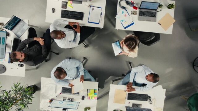 Professional staff use laptops and computers from an overhead angle in a shared office, using digital reports and charts to analyze information for office decisions.