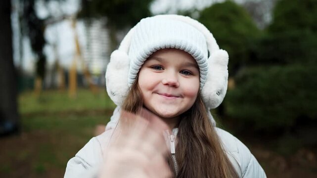 Smiling child in white beanie and fluffy earmuffs reaching toward camera in outdoor park during cool weather, with child, girl, smile, portrait detail, showing winter, beanie, earmuff, jacket context