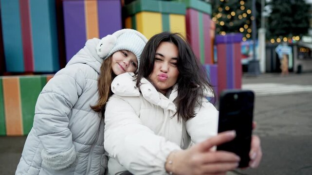 Mother and daughter posing for winter selfie beside oversized colorful gift display in outdoor plaza, smiling in cozy jackets, hats, and earmuffs during festive seasonal celebration