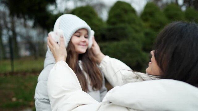 Smiling child in white knit beanie and fluffy earmuffs while adult adjusts winter outerwear in over shoulder park view during soft overcast daylight, with earmuff, beanie, child, girl detail