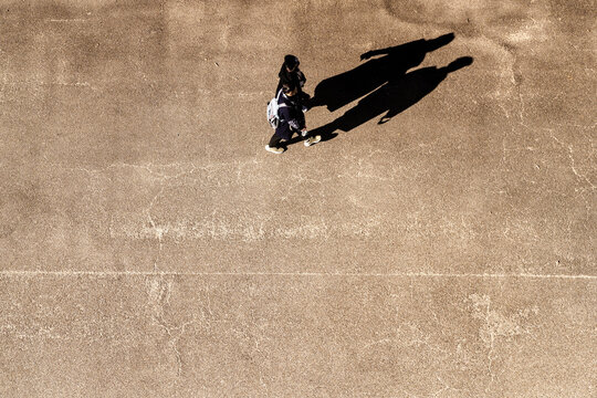 Aerial top down view of a young couple walking closely side by side, their bodies casting one single merged shadow on the pavement on a sunlit park plaza in Japan, high angle city scene