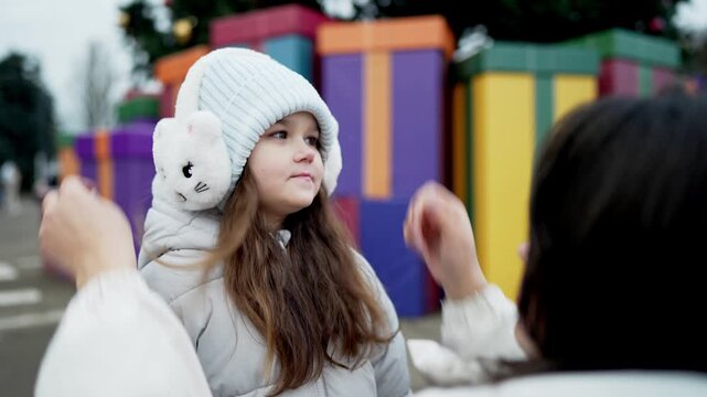 Adult adjusting plush earmuffs and knit hat on child beside oversized gift boxes, decorated evergreen trees, and glowing holiday lights at outdoor winter celebration