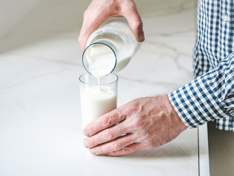 Man's hand pours fresh milk from a glass bottle into a clear glass on a white marble table. Symbolizes dairy nutrition, healthy breakfast, and organic farming. Ideal for food and wellness content