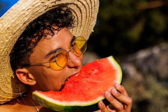 Young man enjoying a sweet watermelon slice wearing a straw hat and yellow sunglasses