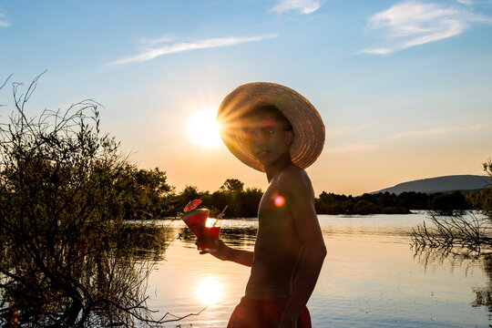 Young man wearing straw hat and sunglasses, enjoying summer vacation by a lake at sunset