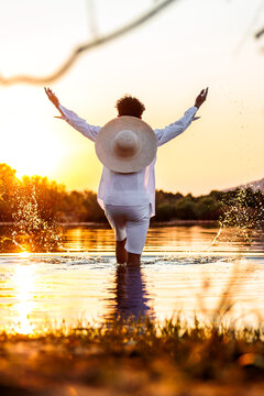 Person with straw hat enjoying freedom at sunset, standing in water and splashing
