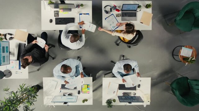 Corporate employees discuss business strategy from an overhead angle in a bright workspace, using market research and planning notes to align objectives for corporate decision making.