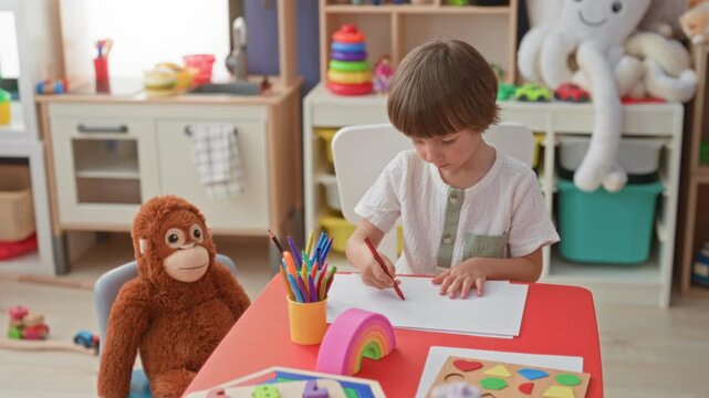 Boy drawing with red pencil at a small red table, hand guiding paper, stuffed monkey seated beside him in building; concentration creativity.