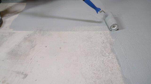 Gray paint being rolled onto concrete floor with blue handled roller. Beige wall white baseboard in background. Partially painted surface shows contrast