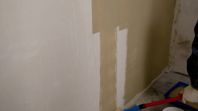 Low angle view of paint roller applying beige paint to wall. Exposed ceiling grid above. Soft lighting highlights texture. Ideal for renovation, home improvement, DIY project visuals