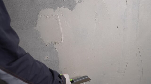 Hand in glove smoothing plaster on wall using trowel. Neutral gray and beige tones. Industrial interior setting. Focus on texture and motion. Ideal for construction, renovation, or DIY content