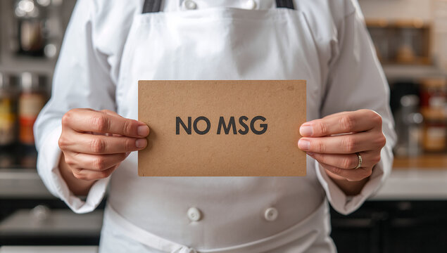 調味料が並ぶオープンキッチンで無添加・化学調味料不使用「NO MSG」カードを掲げるシェフ