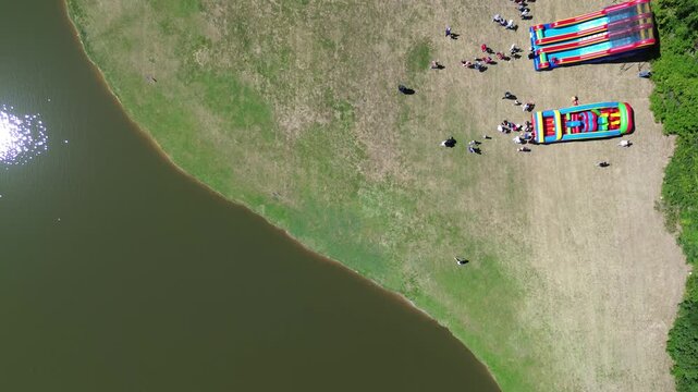 High angle perspective highlights inflatable slides positioned on park grass near shoreline. Long shadows stretch across manicured turf, while dense forest borders open space, Flower Mound, TX