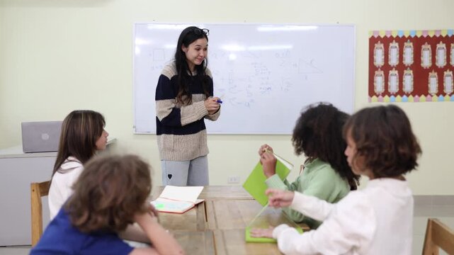 A young female teacher holds a digital tablet and marker while enthusiastically explaining a math lesson to a diverse group of elementary students in a modern classroom.