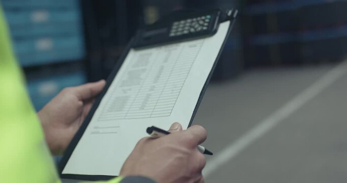 A warehouse worker in a high-visibility vest reviews and notes information on a clipboard in a warehouse aisle.