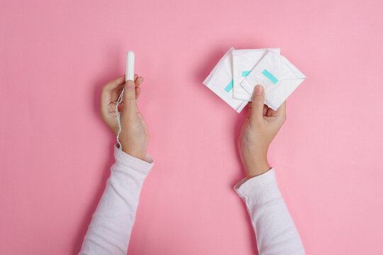Person Holds Tampon In Hand And Three Wrapped Sanitary Pads Isolated On Pink Background