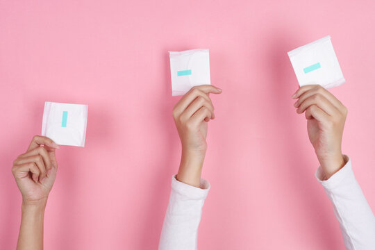 Three Hands Reach Upward Holding Wrapped Sanitary Pads Against Pink Background