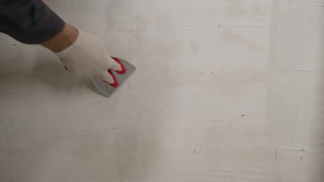 Hand in white glove uses metal trowel to smooth plaster on wall. Motion flows from left to right showing skilled manual labor. Neutral tones dominate