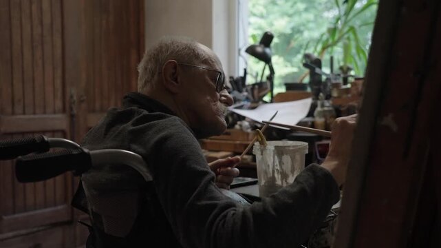 Senior artist on a wheelchair paints an oil painting on canvas in a studio