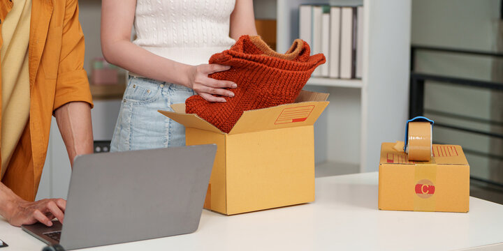 E-commerce business partners packing clothing order and using laptop for shipping