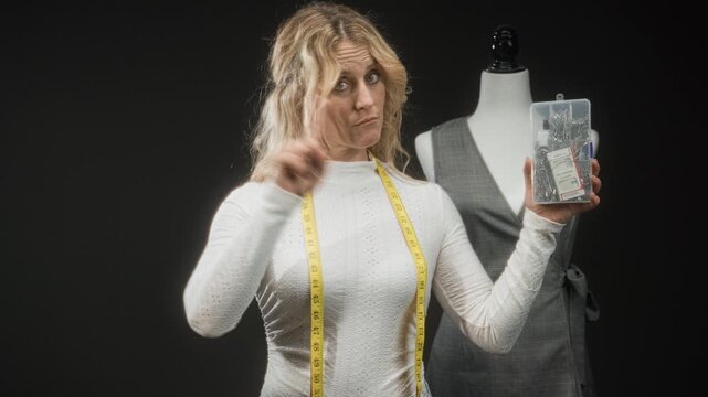 Woman dressmaker with yellow measuring tape holds a clear sewing kit of pins and thread and points finger in a studio beside a mannequin; assertive instruction.