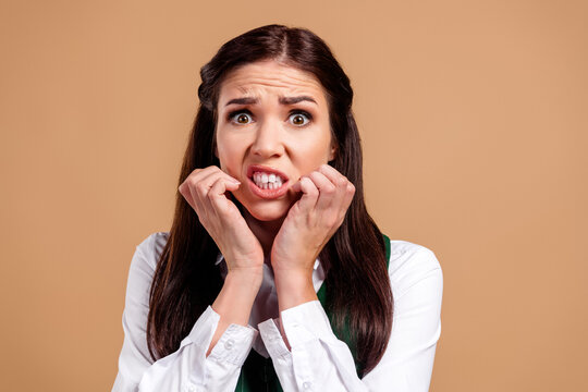 Anxious young woman biting her nails with worried expression and stressed body language, nervousness and anxiety concept portrait on a neutral beige background