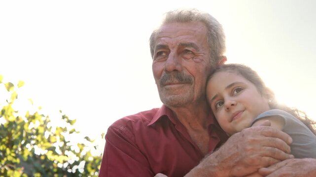 An elderly man embraces a young girl in a sunlit garden as they smile at each other, showing grandparent love, family warmth, trust, tenderness and a peaceful intergenerational bond.