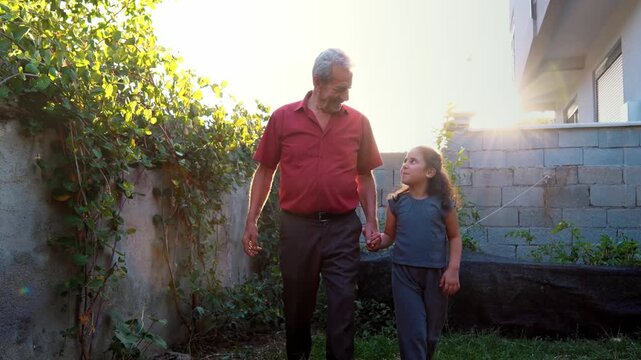An elderly man and young girl walk hand in hand in a green garden, sharing a warm family moment of trust, affection, childhood, nature, intergenerational bond and peaceful rural life.