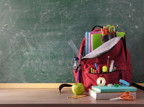 A student school backpack overflowing with colorful stationery supplies placed on a wooden desk in front of a rustic green chalkboard, capturing the nostalgic and inspirational atmosphere of back to