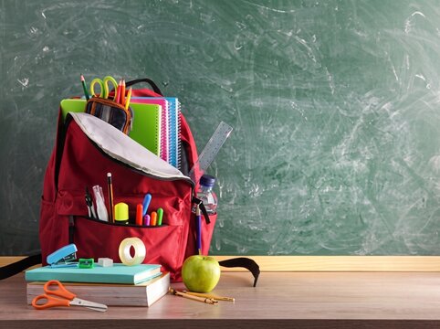 A student backpack filled with essential school supplies sits on a wooden desk before a classic green chalkboard, capturing the academic mood of the back to school season and learning preparation