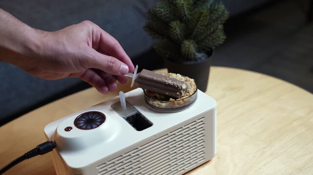 a hand holding a freshly made chocolate ice pop coated in crumbs over a white mini ice pop maker appliance. The device sits on a wooden table in a modern indoor home setting.