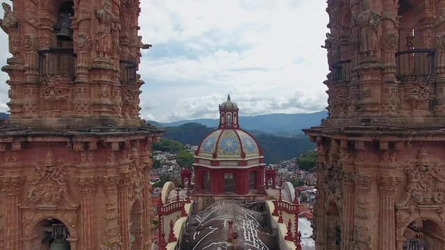Aerial drone view flying between the twin towers of Santa Prisca Church to reveal the colorful tiled dome and colonial town of Taxco, Guerrero, Mexico.