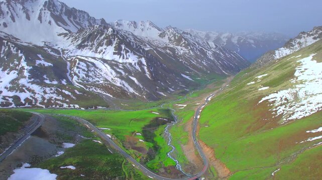 A vehicle driving along a winding mountain road flanked by towering snow-capped peaks; beautiful scenery in Xinjiang, China&mdash;the Duku Highway.