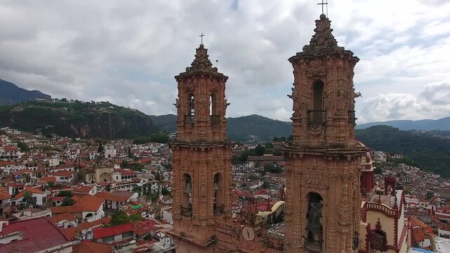 Aerial drone view flying between the twin towers of Santa Prisca Church to reveal the colorful tiled dome and colonial town of Taxco, Guerrero, Mexico.