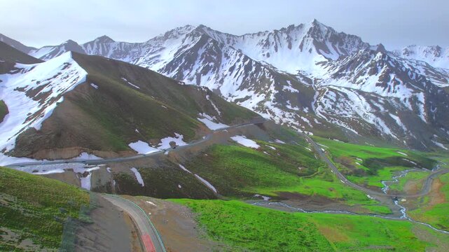 A vehicle driving along a winding mountain road flanked by towering snow-capped peaks; beautiful scenery in Xinjiang, China&mdash;the Duku Highway.