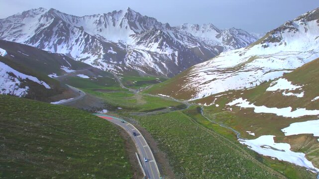 A vehicle driving along a winding mountain road flanked by towering snow-capped peaks; beautiful scenery in Xinjiang, China&mdash;the Duku Highway.