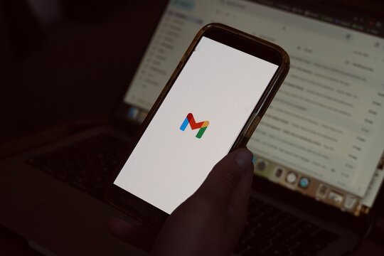 Barcelona, Spain - March 19, 2026: A person holds a smartphone displaying the Gmail logo and app loading screen, with a laptop showing an email inbox in the background
