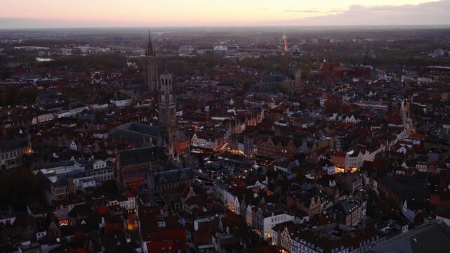 Drone footage starting with a wide overview of Bruges and moving towards the Grote Markt, showing the historic city center, evening lighting, and illuminated buildings.
