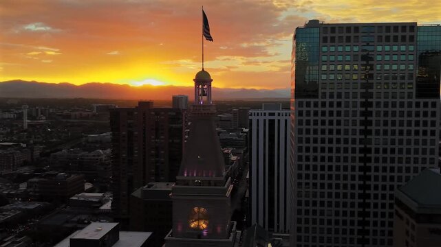 Closer orbit around the top of the Daniels and Fisher Tower and flag, moving right to reveal a glowing sunset and the Rocky Mountain skyline.