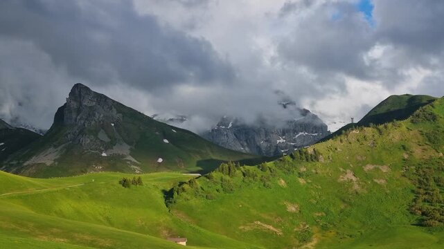 Dramatic summer view of the Swiss Alps in Adelboden, Lenk, Switzerland, with towering mountain peaks beneath a stormy sky. Rolling clouds cast striking shadows across the green alpine landscape