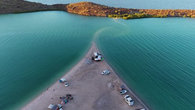 A compelling high-angle drone sequence flying over camper vans parked at the tip of a narrow sand spit where it disappears into the turquoise bay.